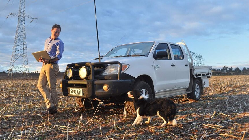 A man with a laptop open stands next to a ute and a dog on a farm.
