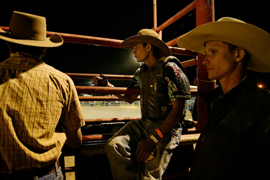 Three men wearing cowboy hats standing next to a fence surrounding a rodeo ring, at night. 