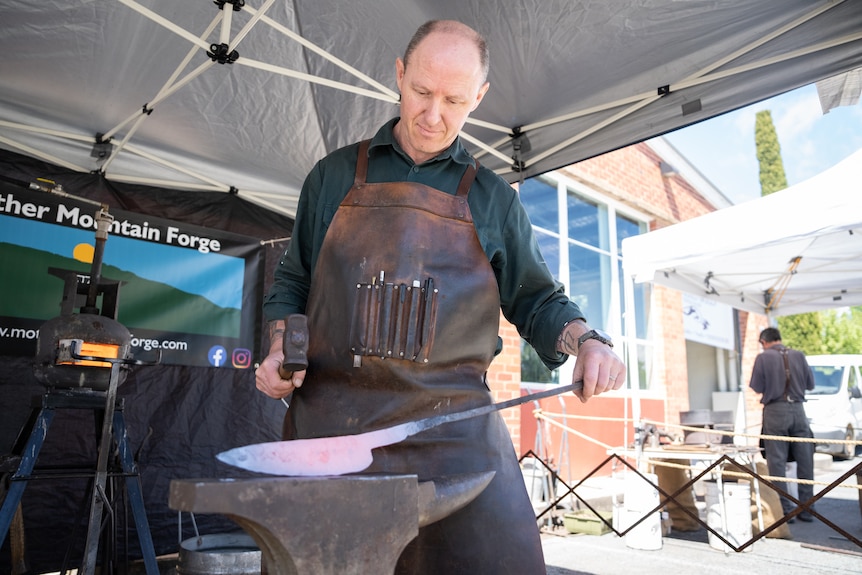 The man wears a leather apron and hammers a big metal knife on a forge.
