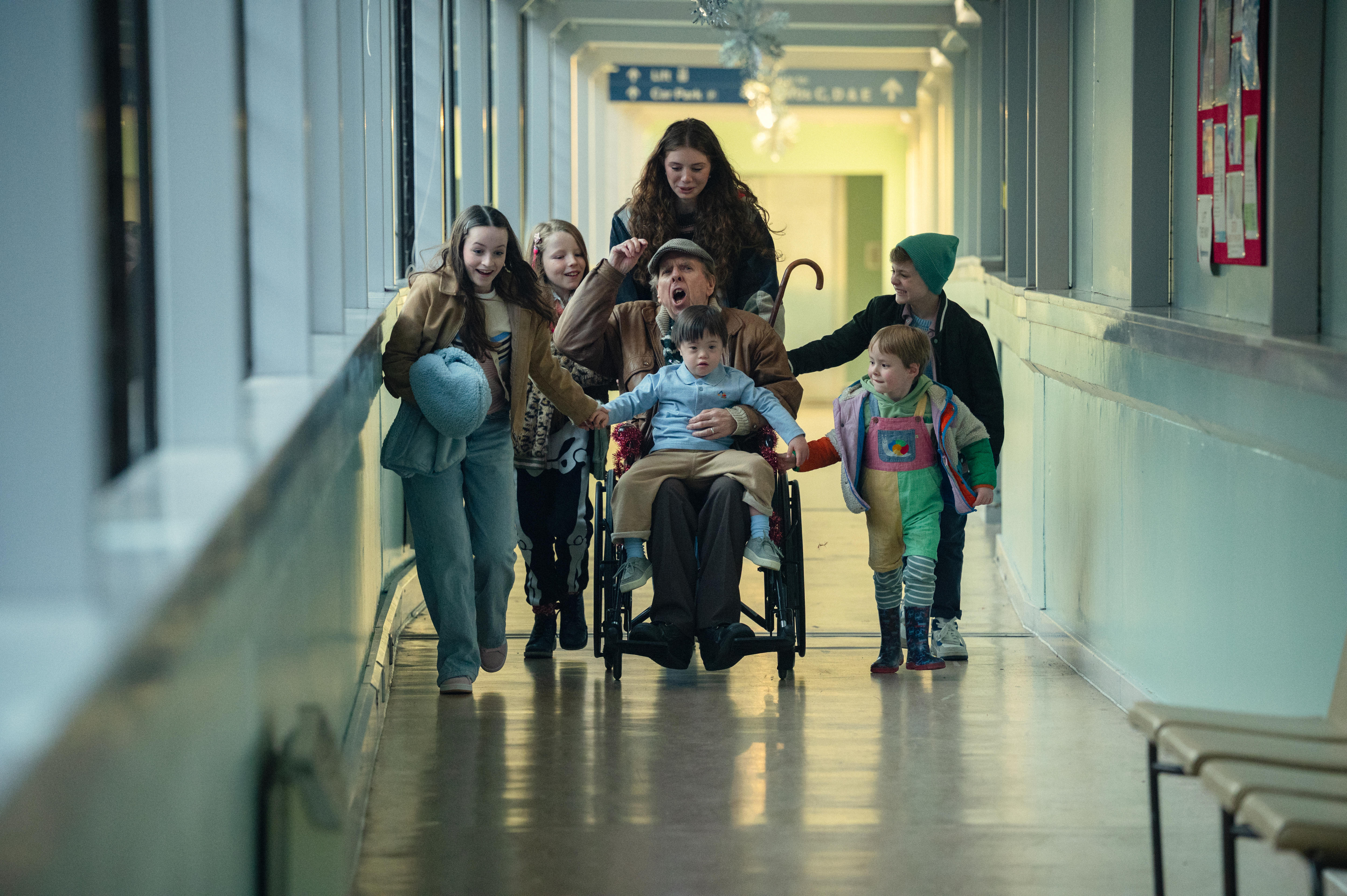 Un grupo de niños empuja a un anciano en silla de ruedas con un bastón por el pasillo de un hospital mientras él vitorea.