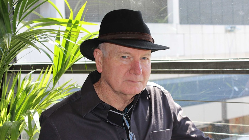 man in hat and brown jacket looks at camera form side with greenery on a balcony behind