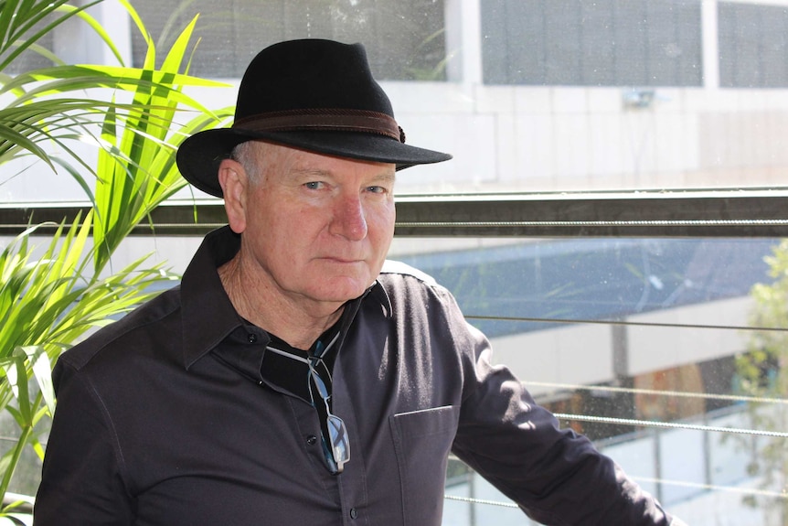 man in hat and brown jacket looks at camera form side with greenery on a balcony behind