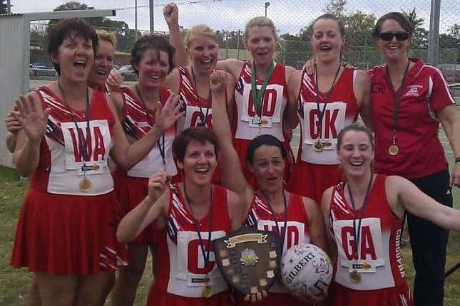 A netball team celebrates winning with medals around their necks and holding a trophy.