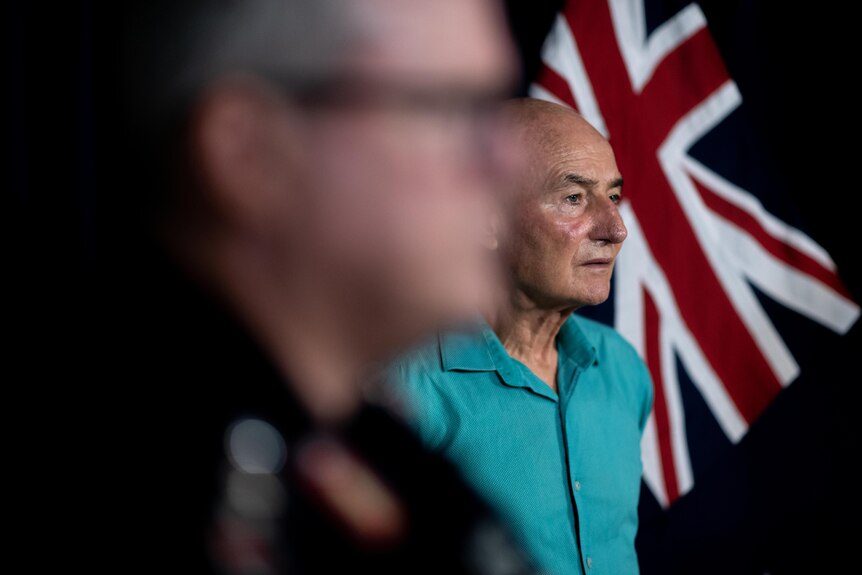 A man wearing a green shirt and looking seriously stands in front of a flag.