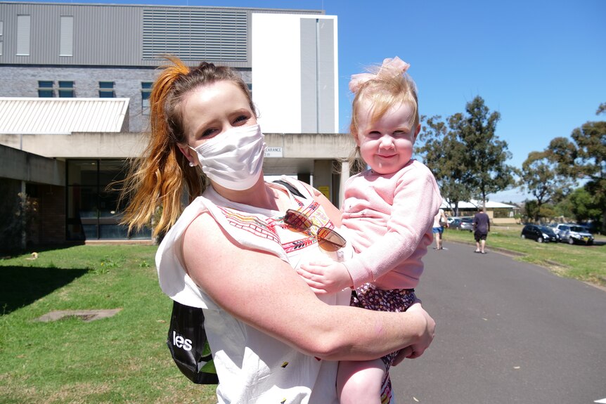 A masked woman holding a small, smiling girl in her arms on a cloudless day.