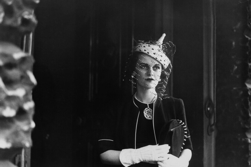 A young woman wearing a dark dress, white gloves and a black fascinator looks off in the distance.