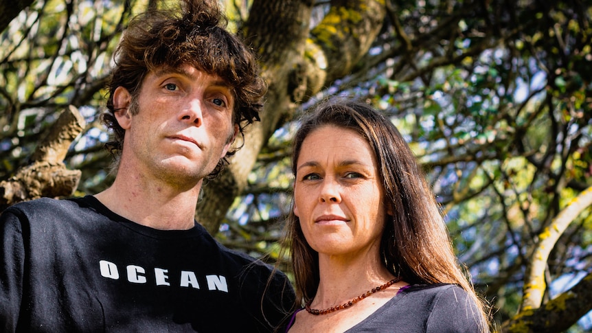 A man and woman stand under a tree on a sunny day looking down at the camera.
