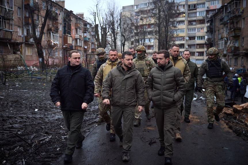 A group of men wearing winter clothing and combat fatigues walk through a street surrounded by heavily damaged buildings.