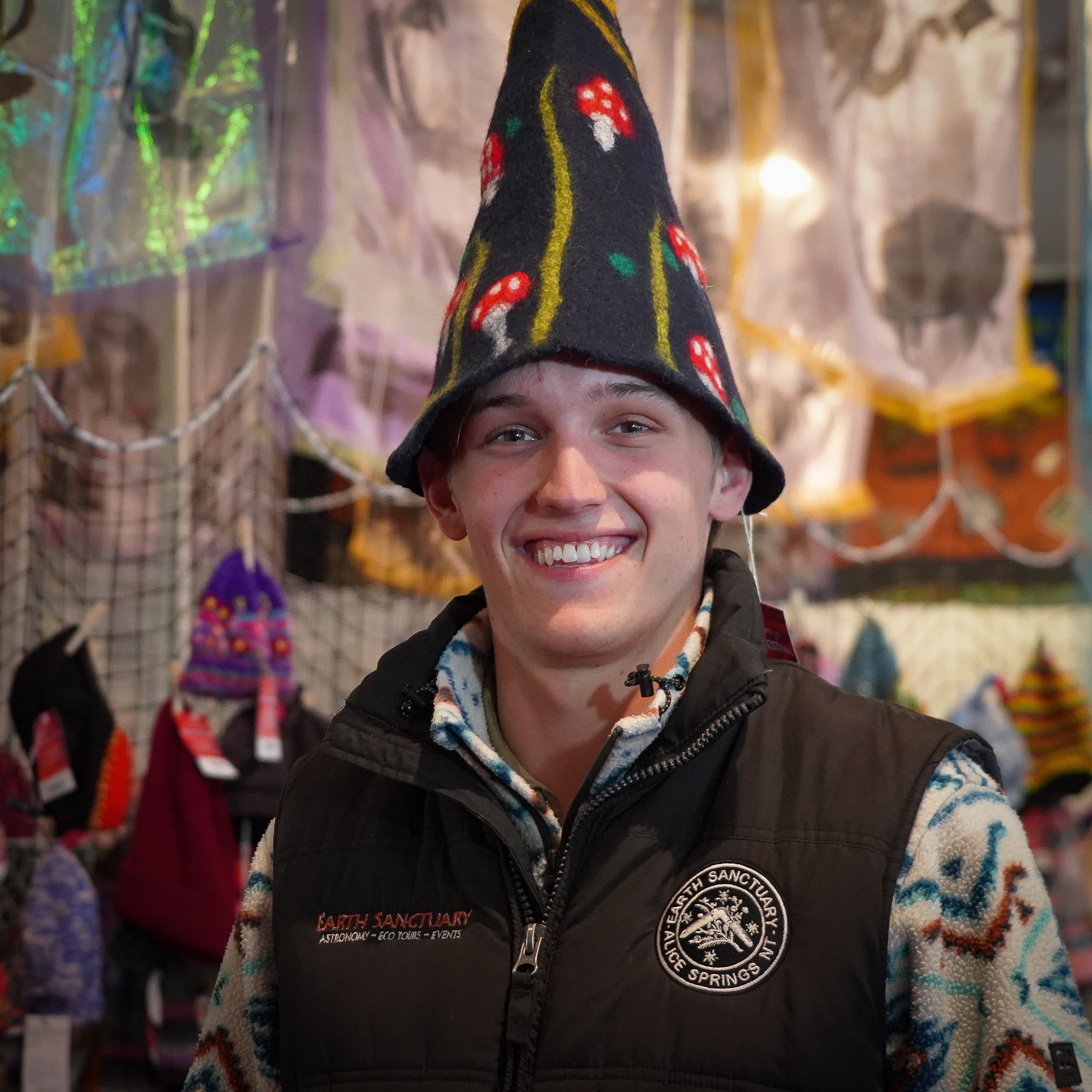 A smiling man wears a pointy felt hat, tall trees and red and white mushrooms on the beanie depict a forest scene.