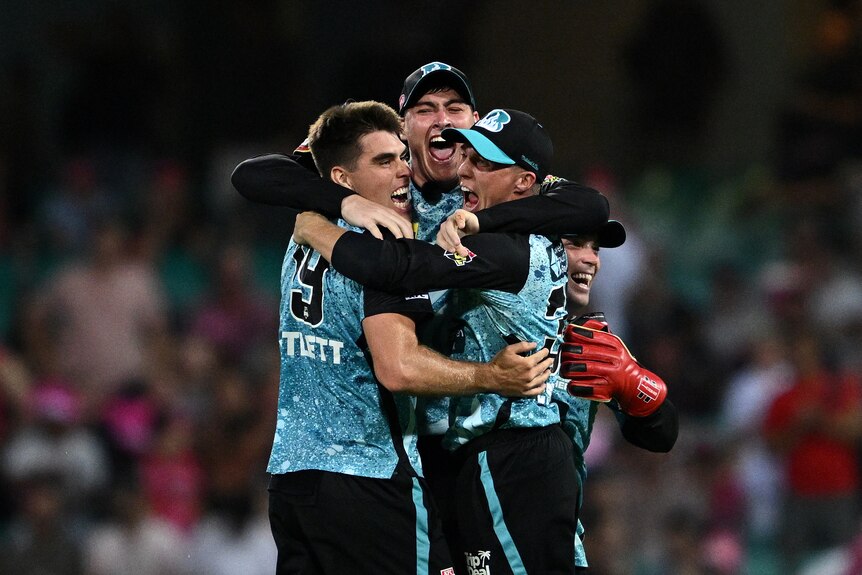 Three men in blue and black cricket uniforms hug and smile.