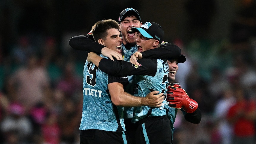 Three men in blue and black cricket uniforms hug and smile.