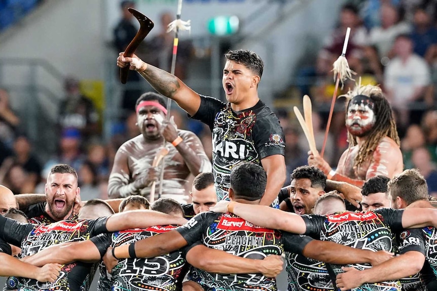 Latrell Mitchell stands and shouts while surrounded by kneeling Indigenous All Stars during a war cry.