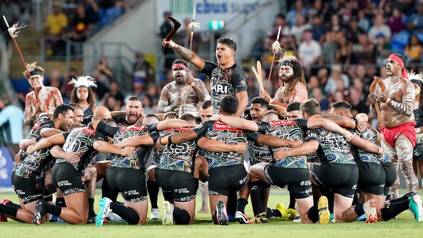 Latrell Mitchell stands and shouts while surrounded by kneeling Indigenous All Stars during a war cry.