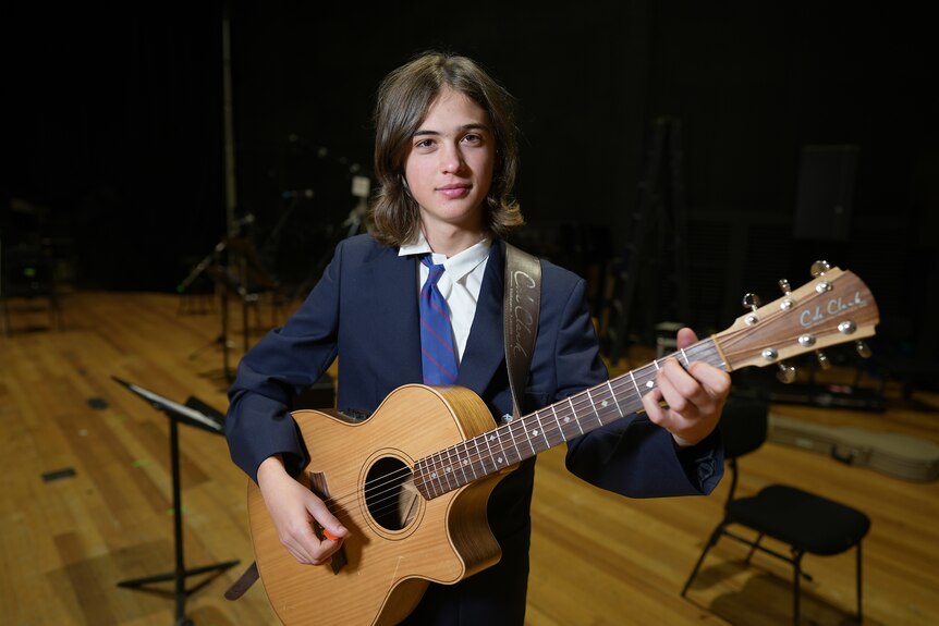 Rory, wearing a school tie and blazer, holds a guitar.