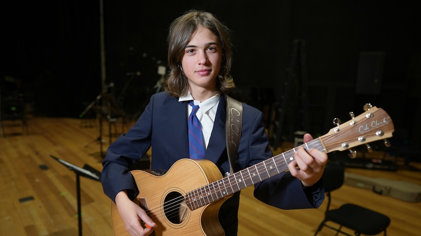 Rory, wearing a school tie and blazer, holds a guitar.