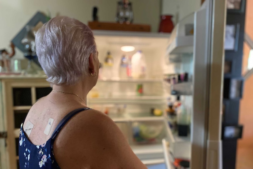 A woman with white hair, in blue top, looks into an open fridge. There are a few items in there, but it is noticeably bear.