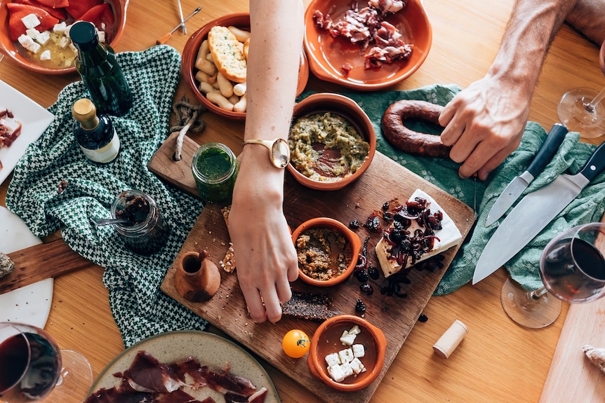 A hand reaches across a table full of cheese, dip, cured meat and other snack foods to pick a snack. 