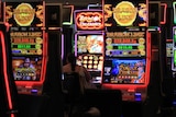 A woman sits at a poker machine. Surrounding her are several large, bright and colourfully lit machines.