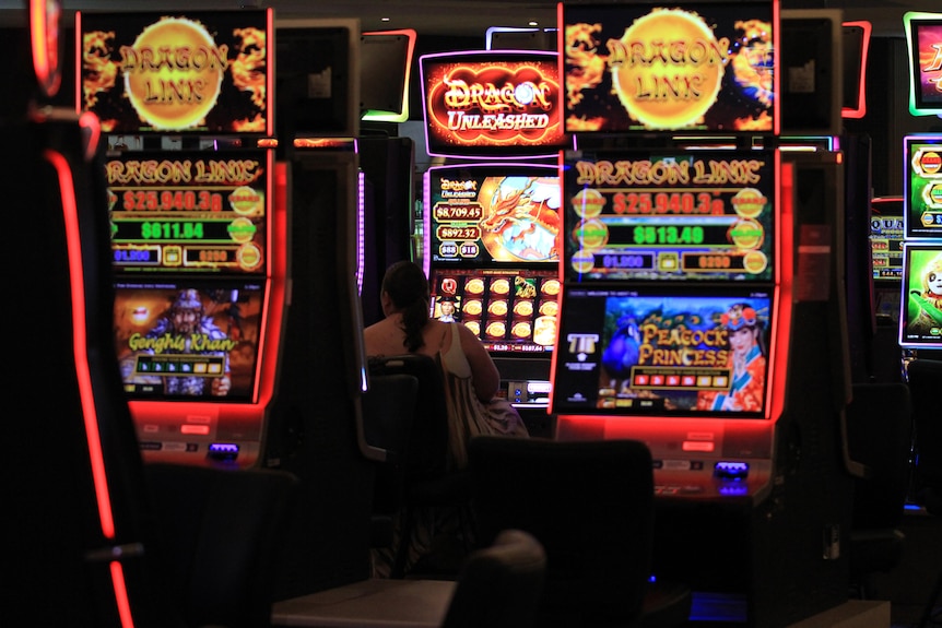 A woman sits at a poker machine. Surrounding her are several large, bright and colourfully lit machines.