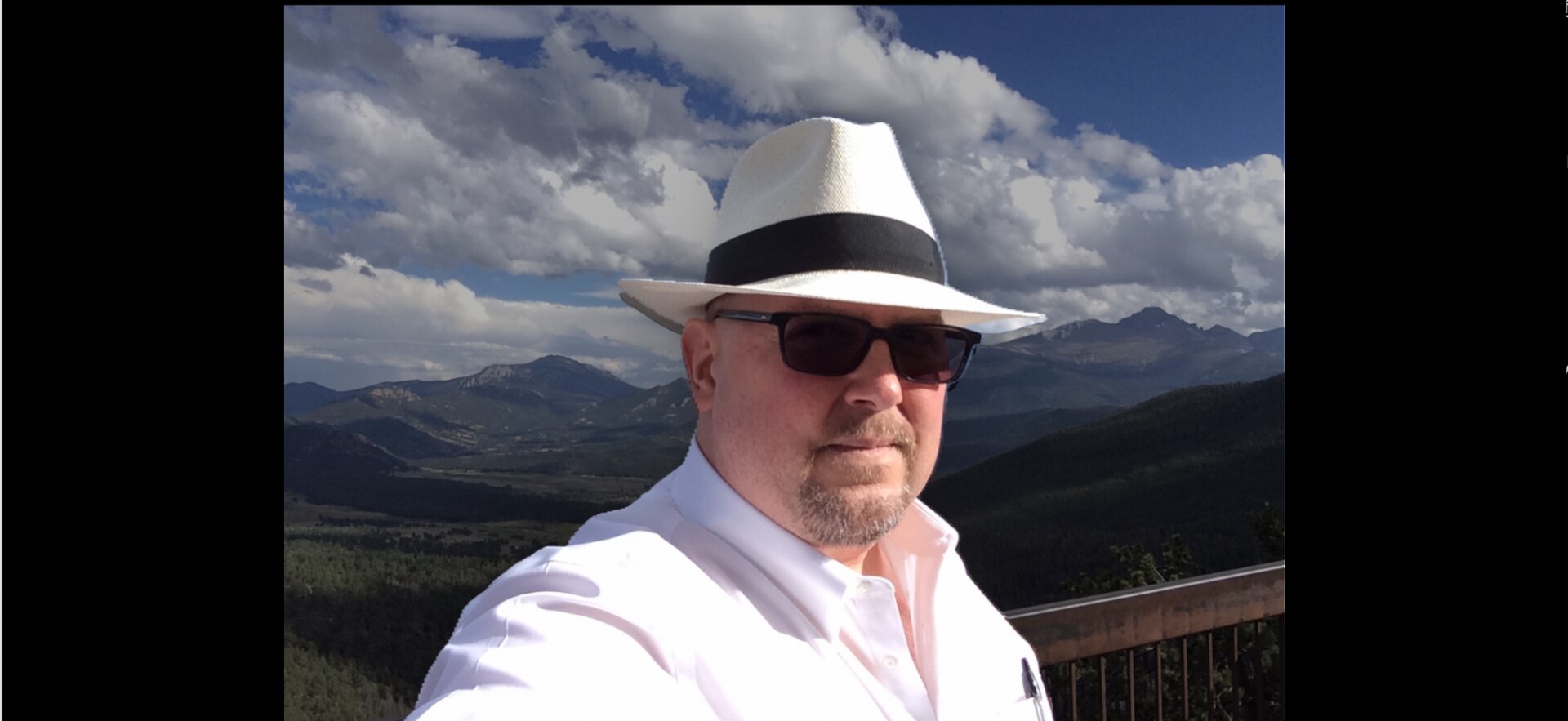 A man in a collared shirt, white panama hat and sunglasses takes a selfie in front of a mountain range