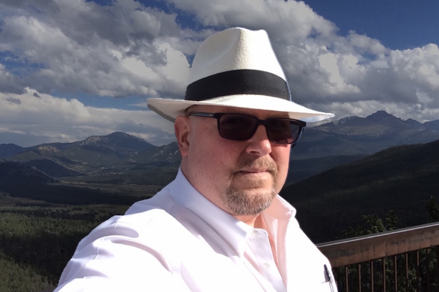 A man in a collared shirt, white panama hat and sunglasses takes a selfie in front of a mountain range