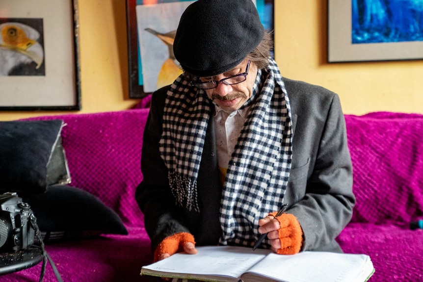 A man sits on a bright purple couch looking at a folder of documents. He has bright orange woollen gloves on.