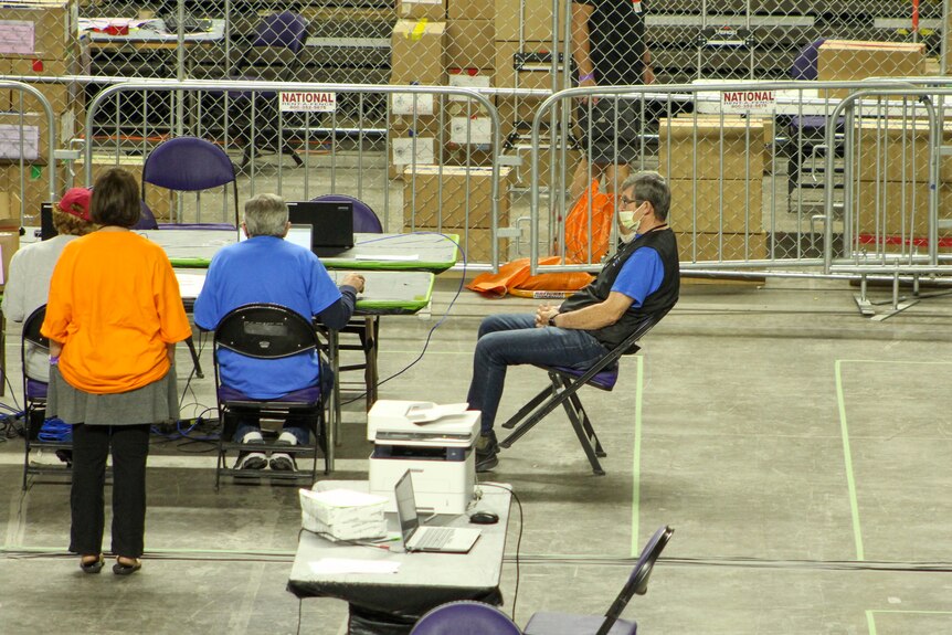 A man in jeans a blue shirt leans back in his chair while others keep counting ballots 