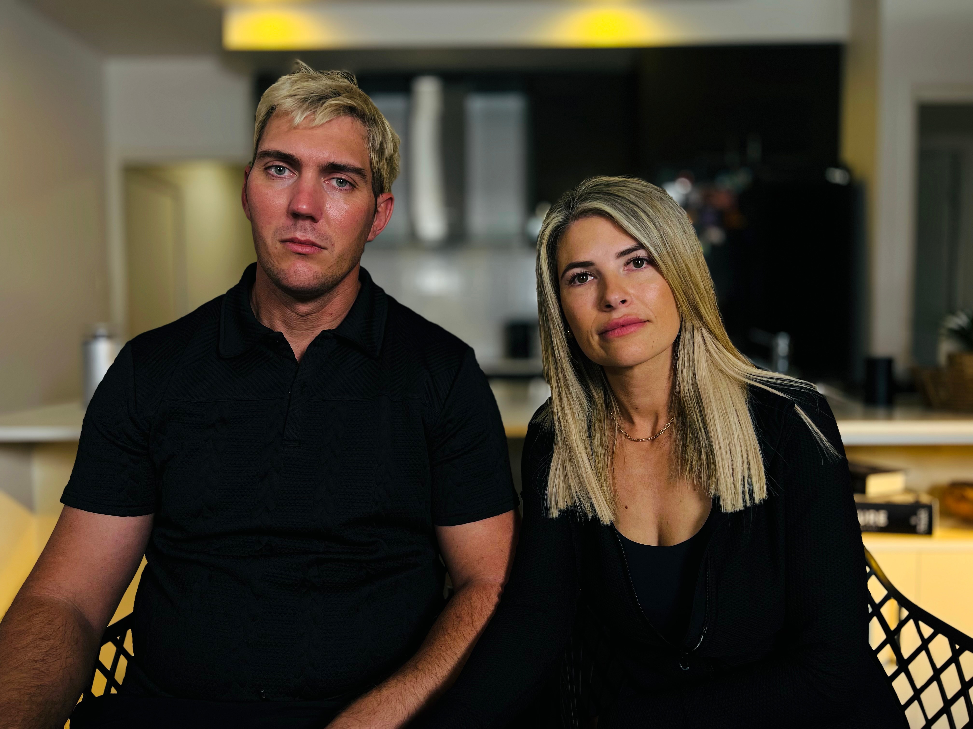 A man and a woman sit in an apartment. They are looking at the camera with serious expressions.