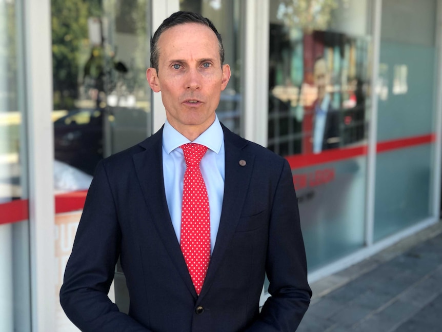 A man with dark hair and blue eyes, wearing a suit with a bright red tie looks into the camera.