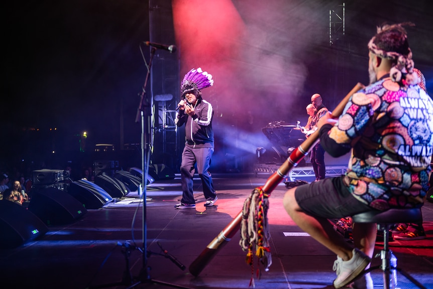 Jay Kay of Jamiroquai on stage with a didgeridoo player who wears a shirt saying 'always was always will be'