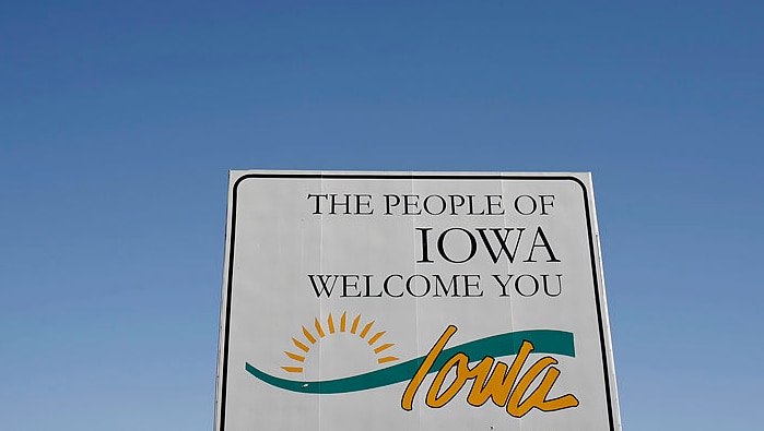A road sign in the foreground with "The People of Iowa Welcome You" and a cloudless blue sky in the background.