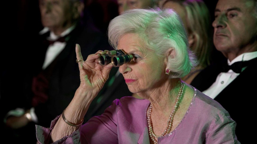 A woman in theatre seats holds binocular opera glasses to her face with a raised little finger.