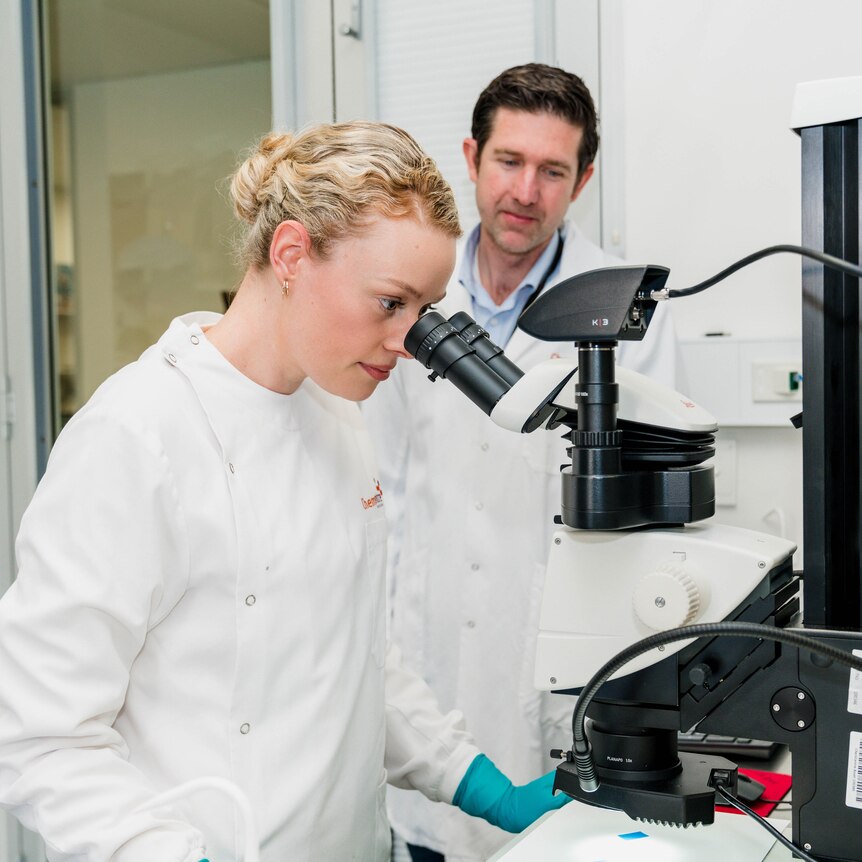 A woman with blonde hair in a white lab coat looks at a microscope 