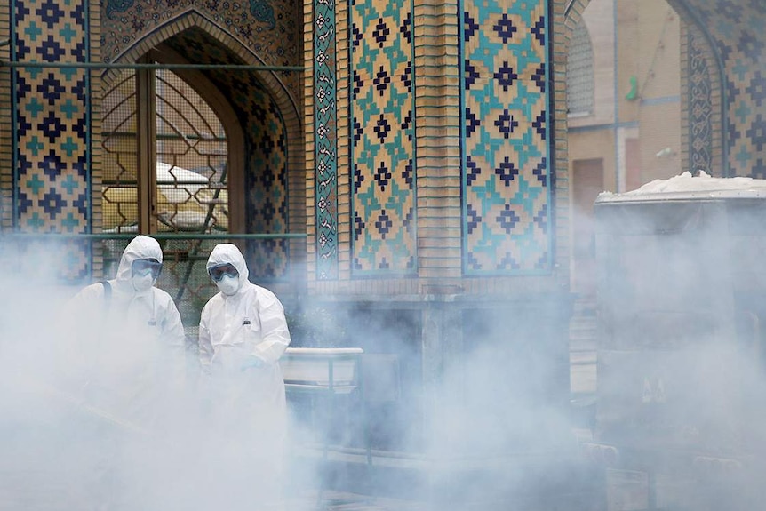 Disinfectant spray rises above workers in hazard suits in front of a religious shrine, in Iran.