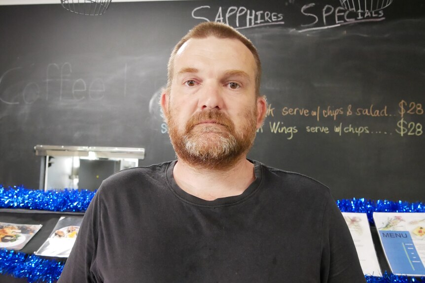 Un homme avec une barbe portant une chemise noire.  Il y a un menu au tableau en arrière-plan.