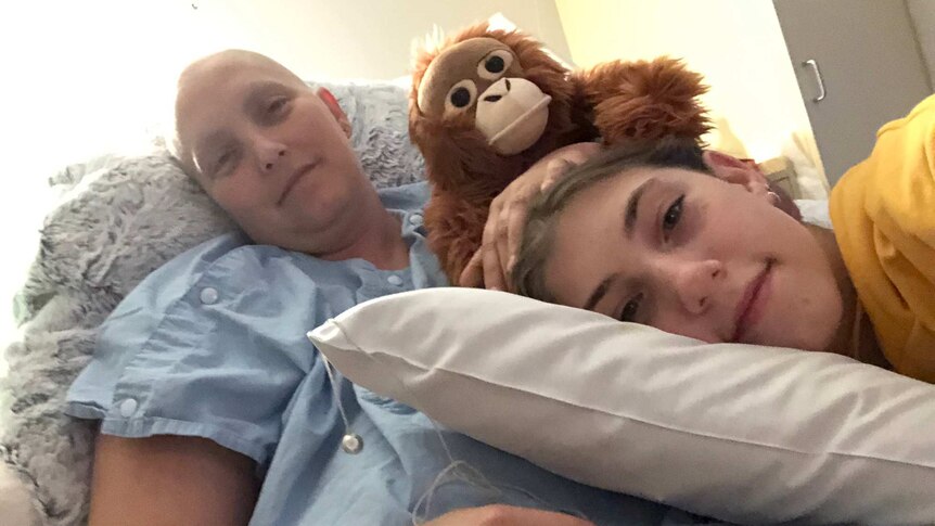 A woman who is bald and undergoing treatment for cancer lies on a bed in hospital with her young daughter lying on her stomach.
