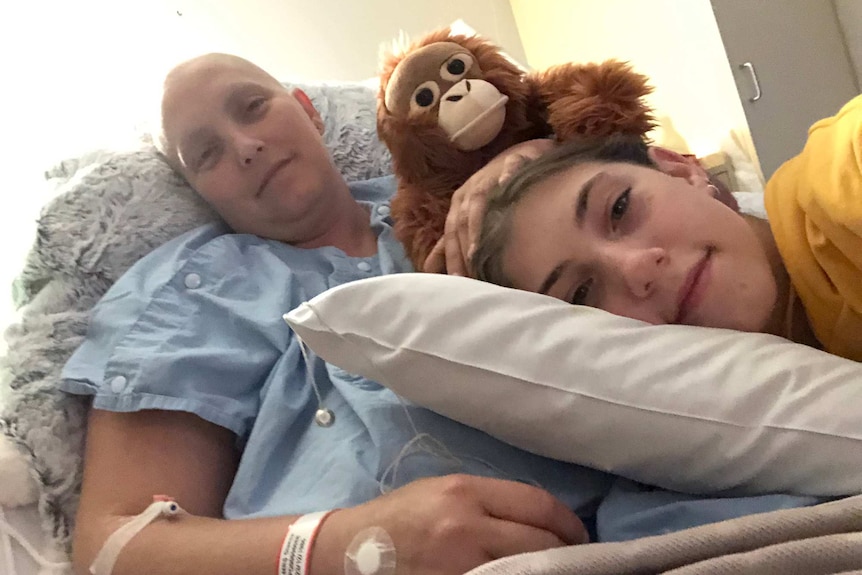 A woman who is bald and undergoing treatment for cancer lies on a bed in hospital with her young daughter lying on her stomach.