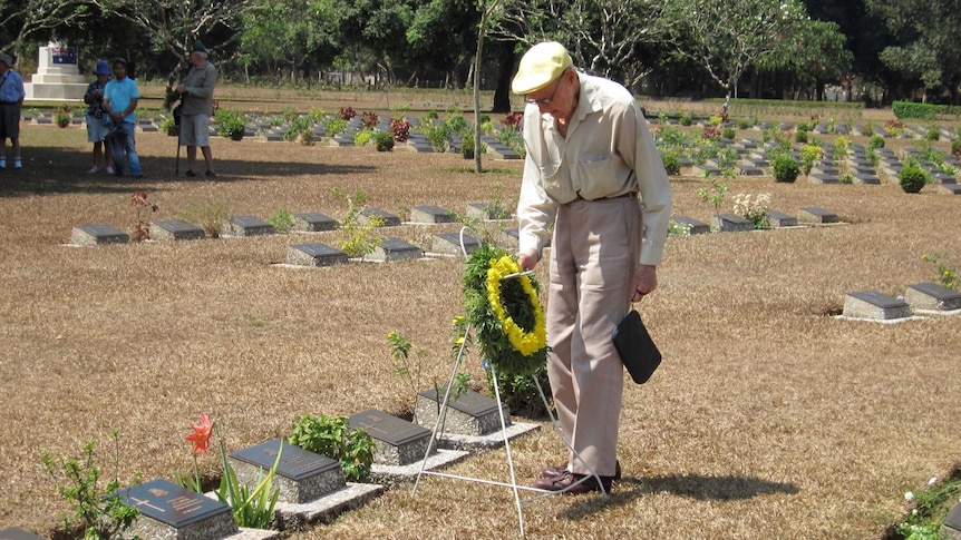 Elderly man wearing flat cap and holding wreath with yellow flowers at cemetery