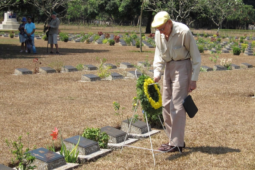 Elderly man wearing flat cap and holding wreath with yellow flowers at cemetery