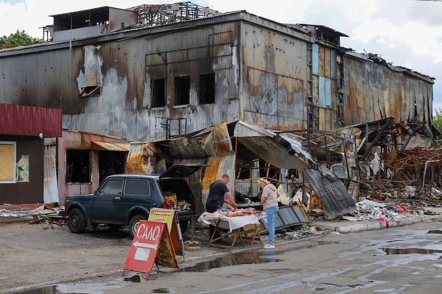 A man selling meat out of the back of his car in front of a destroyed building.