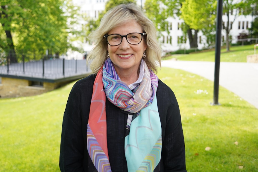 A woman wearing glasses and a scarf stands on a manicured lawn.