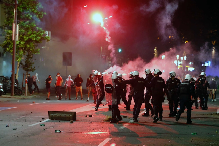 A flare lights up the night sky above  protesters and a group of police in riot gear.