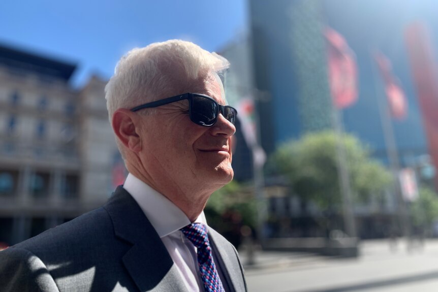 A man with white hair, wearing sunglasses and a suit, standing in sunshine.