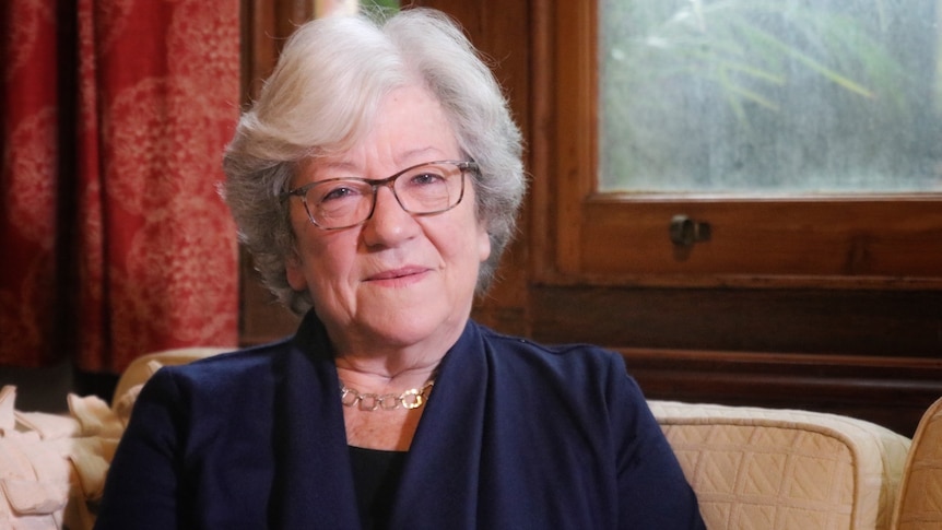 An older, bespectacled woman – former WA premier Carmen Lawrence – smiling faintly as she sits in a sitting room.