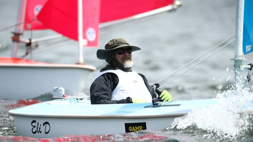 A man with a short grey beard, sunglasses and a broad brimmed hat smiles as he sails a one-person yacht.