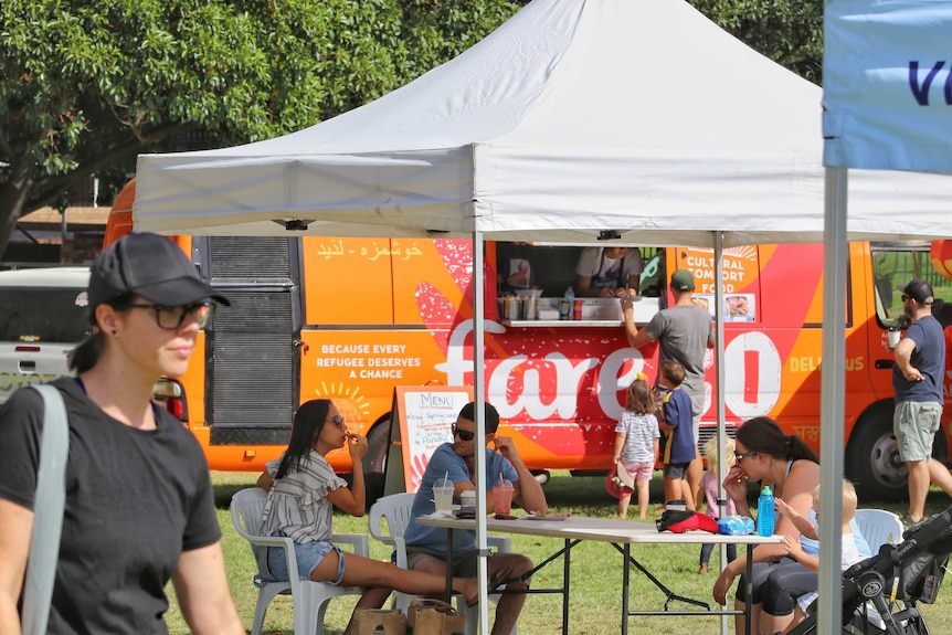 A woman walks in front of a group of people sitting down at a farmers market with a food truck in the background.