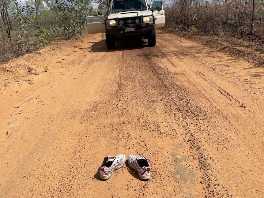 A pair of sneakers sits in the middle of a dirt track with the front of a four-wheel-drive vehicle in the shot.