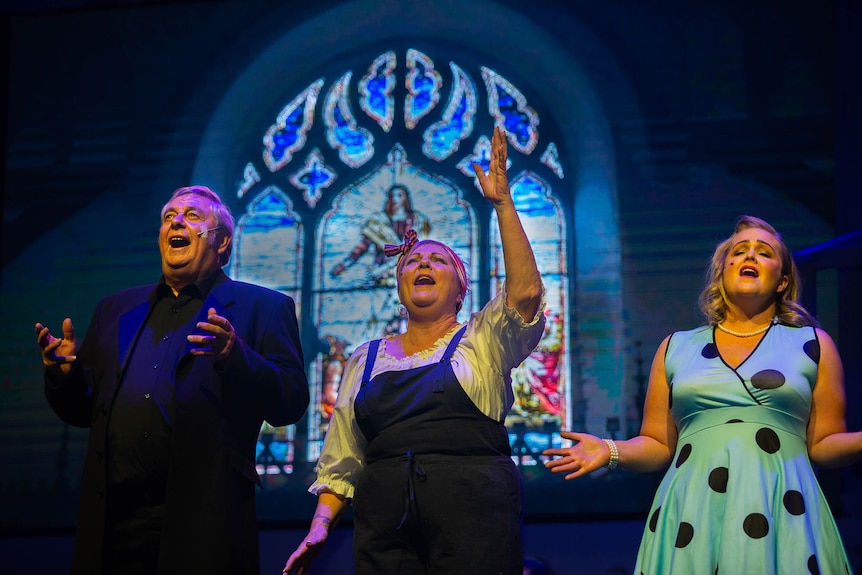 Three singers look towards the roof, singing in front of projected church stained glass.