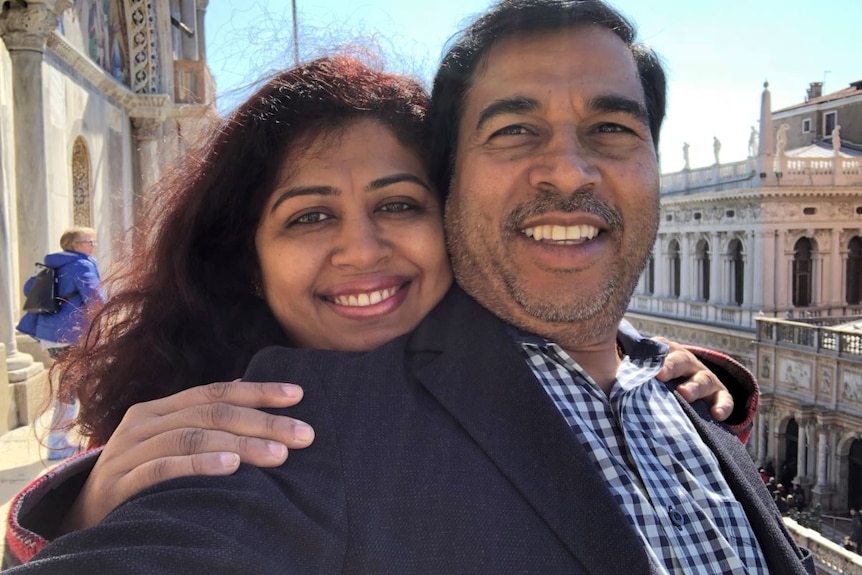 A woman stands behind a man and puts her hand on his shoulders as they both smile at the camera for a selfie on a terrace.