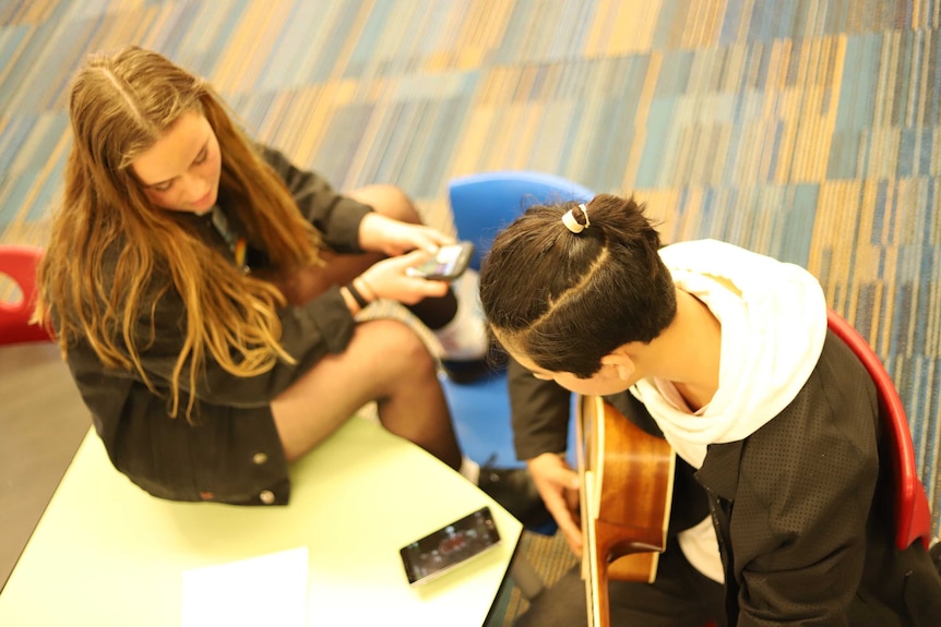 Guy and girl sitting on the carpet looking at phones in classroom.